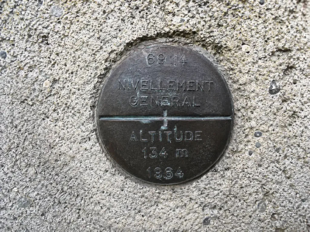 Gros plan réaliste d’un repère de nivellement en métal sombre scellé dans un mur en béton granuleux, avec les inscriptions “NIVELLEMENT GÉNÉRAL”, “ALTITUDE 134 m” et “1964”.